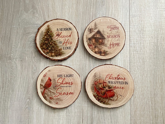 Set of four rustic wood slice coasters featuring faith-based winter scenes including a cardinal, sleigh, Christmas tree, and log cabin with inspirational messages.