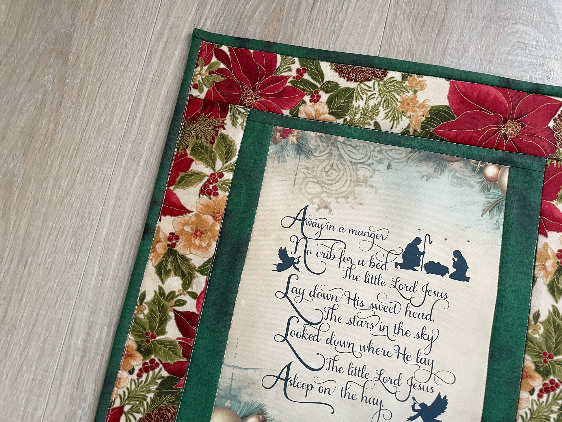 Handmade quilted Christmas wall hanging with an “Away in a Manger” verse framed by red poinsettias.
