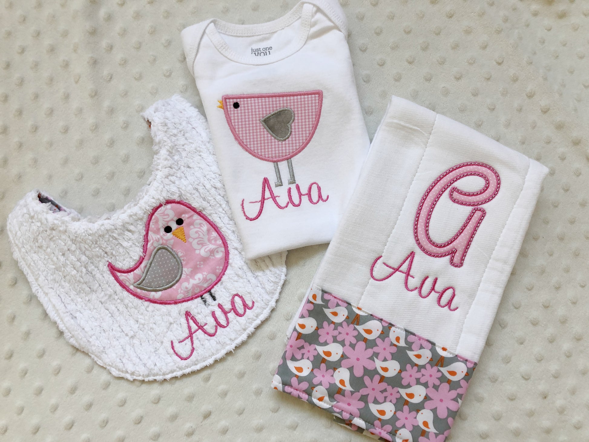 A personalized 3-piece baby girl gift set featuring a pink bird bodysuit, a white chenille bib, and a monogrammed burp cloth.