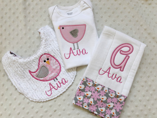 A personalized 3-piece baby girl gift set featuring a pink bird bodysuit, a white chenille bib, and a monogrammed burp cloth.