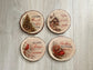 Set of four rustic wood slice coasters featuring faith-based winter scenes including a cardinal, sleigh, Christmas tree, and log cabin with inspirational messages.