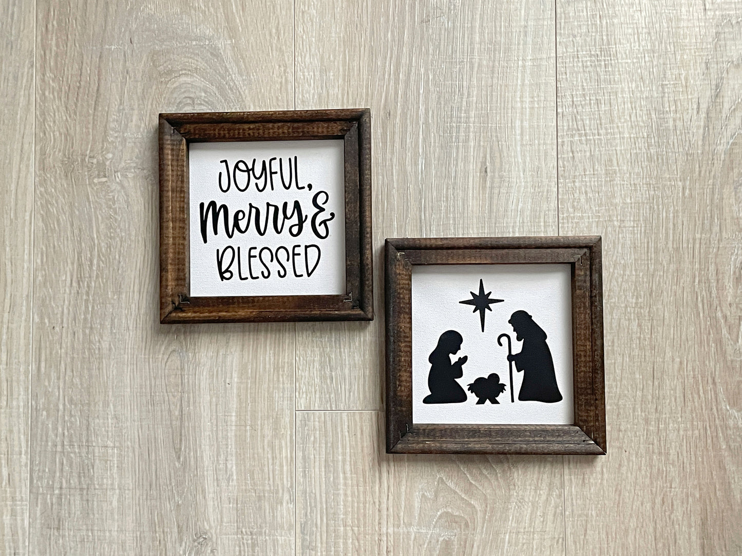 Two wood-framed Christmas signs featuring a nativity silhouette and the phrase "Joyful, Merry & Blessed" on soft white backgrounds.