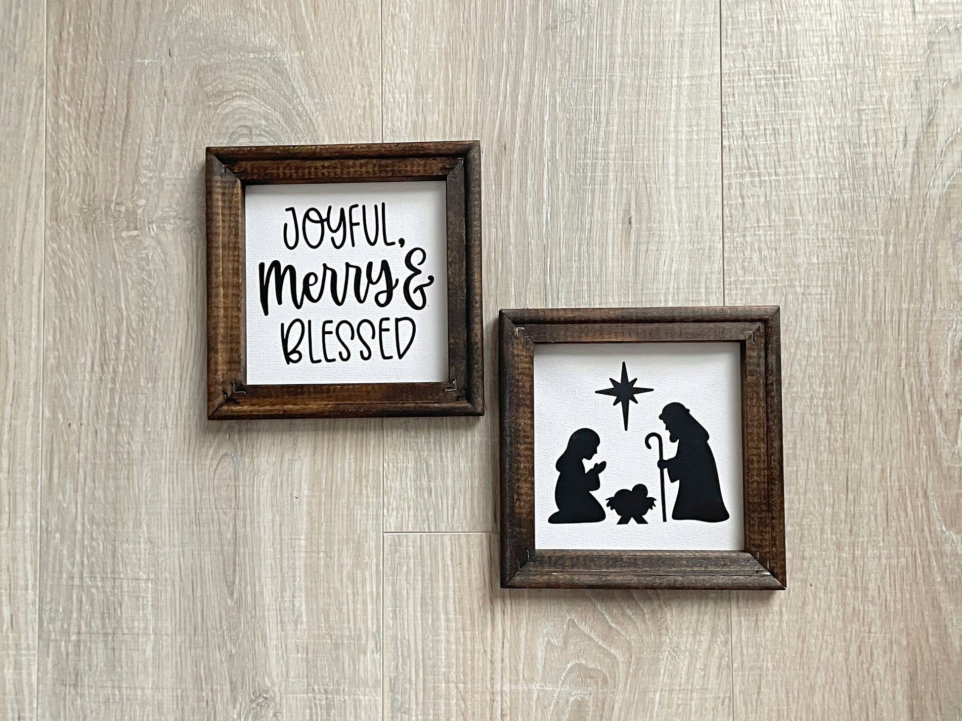 Two wood-framed Christmas signs featuring a nativity silhouette and the phrase "Joyful, Merry & Blessed" on soft white backgrounds.