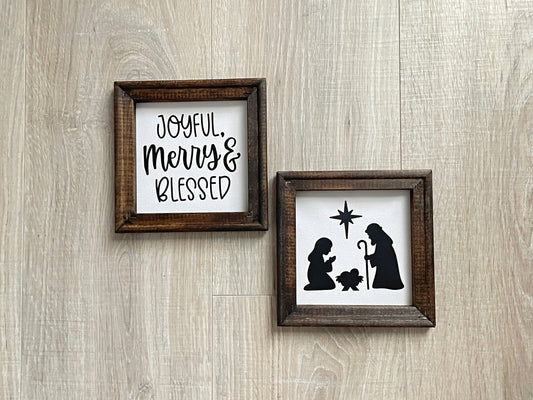Two wood-framed Christmas signs featuring a nativity silhouette and the phrase "Joyful, Merry & Blessed" on soft white backgrounds.