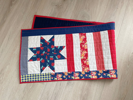 A 15x52 inch quilted patriotic table runner featuring a blue floral star pattern and red and white patchwork stripes.
