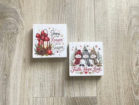 Two freestanding winter shelf sitters featuring "Jesus Is the Reason for the Season" and "Faith Hope Love" designs with snowmen and holiday accents.
