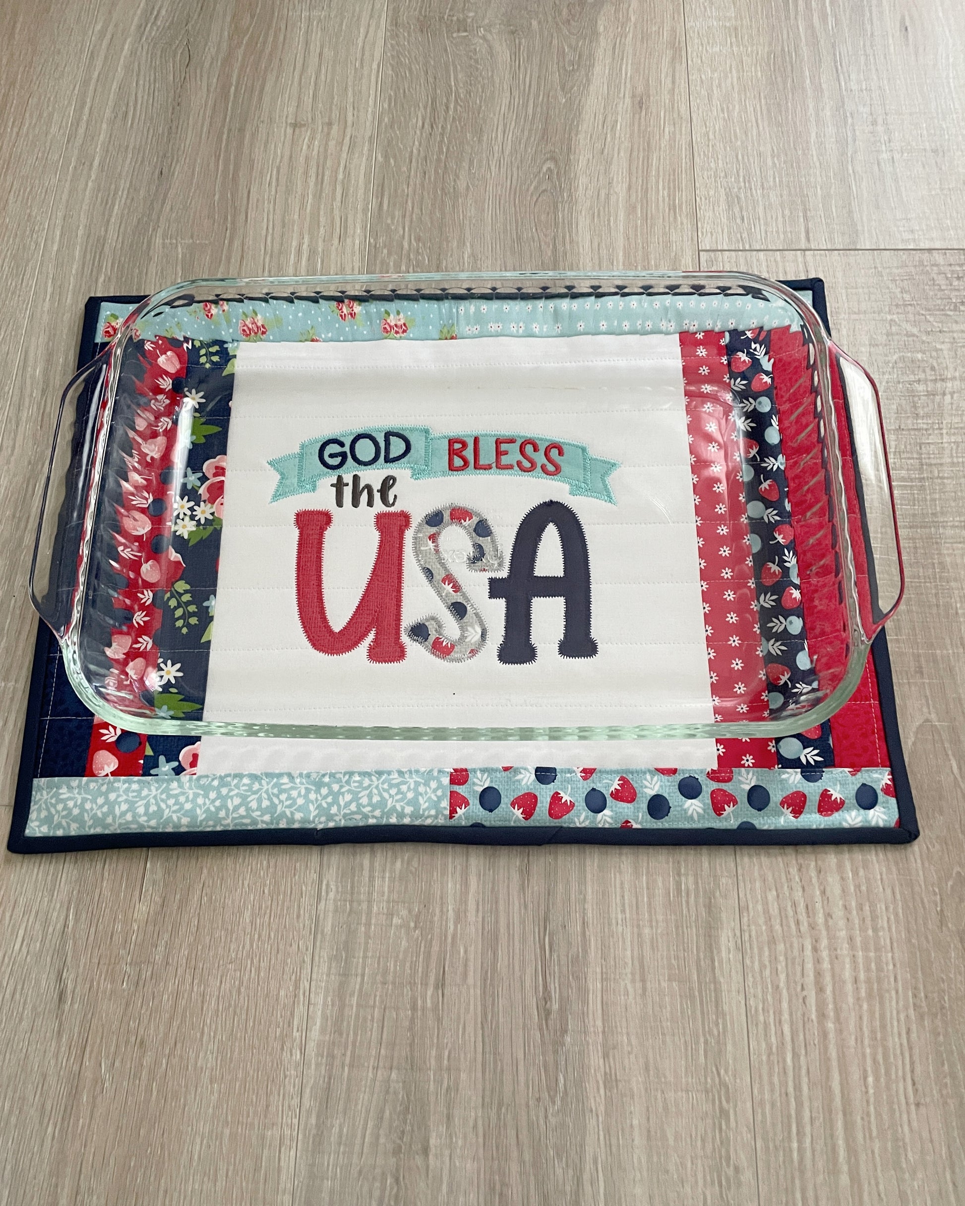 This 12 x 16 red, white, and blue quilted casserole hot pad/trivet has a scrappy patchwork center panel featuring a machine embroidered design -- God Bless the USA. These are perfect for a 9 x 13 casserole dish or 2 smaller baking dishes. Shown underneath a 9 x 13 casserole dish.